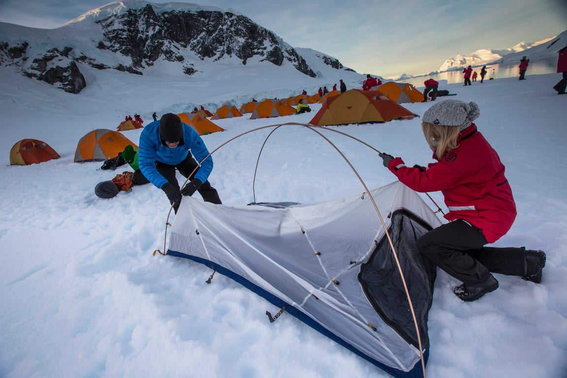 Expedition travelers setting up camping tents on Antarctic ice with mountains in background